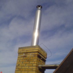 ZAKŁAD URZĄDZEŃ GRZEJNYCH I ŚLUSARSTWA ANDRZEJ KREFT w spadku - Komin z cegły z metalowym wkładem kominowym, widoczny fragment dachu, na tle błękitnego nieba z chmurami.