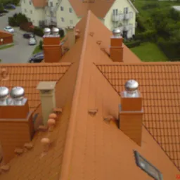 Czerwony dach wielospadowy z kominami, wentylatorami turbinowymi i oknami dachowymi, widok z góry na tle zabudowy mieszkalnej i zieleni.