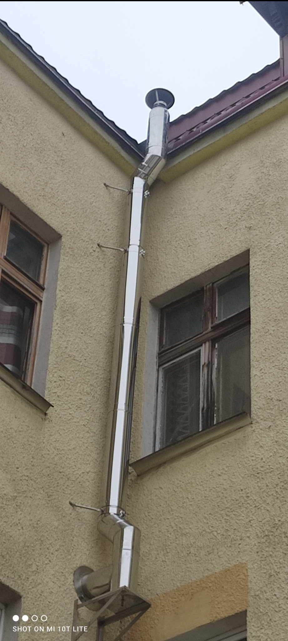 Zewnętrzny, pionowy, metalowy przewód kominowy zamocowany do ściany budynku, widoczny z boku, z uchwytami mocującymi, obok okna z drewnianą ramą.