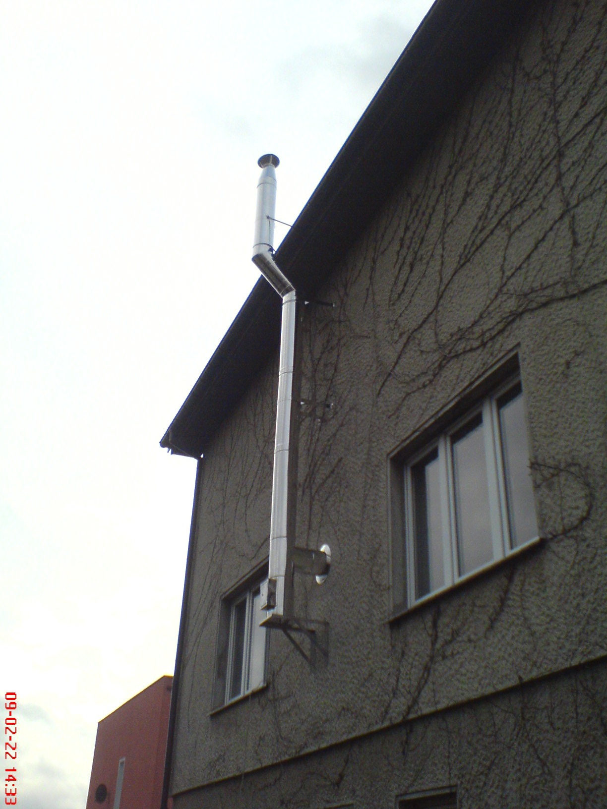 Zewnętrzny, stalowy komin dymowy przymocowany do ściany budynku z szorstkim tynkiem, porośniętej suchym bluszczem, widoczny fragment dachu i okna.