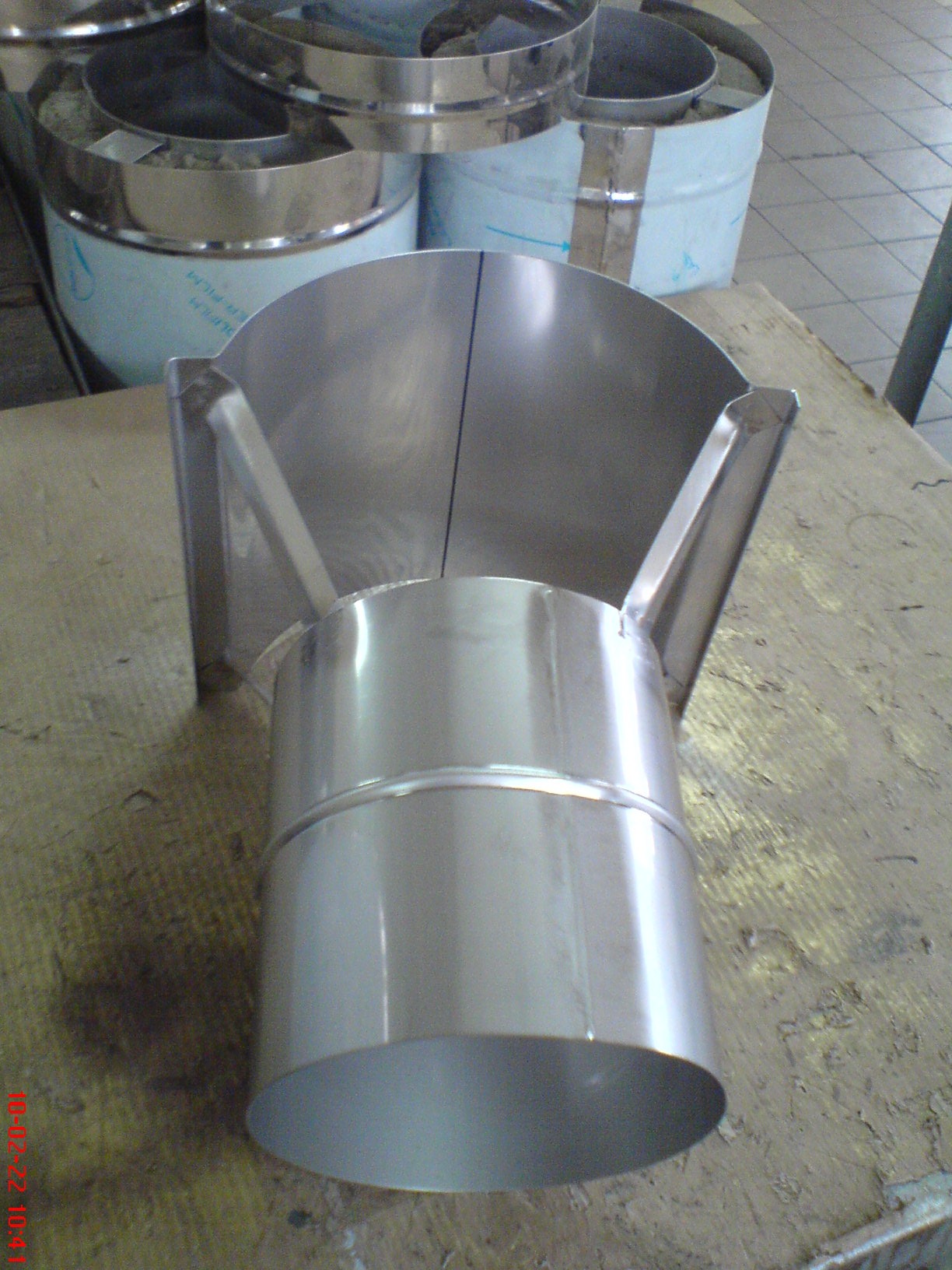 Metalowy element wentylacyjny z blachy nierdzewnej, widoczne spawane łączenia, na tle inne elementy wentylacyjne i podłoga wyłożona płytkami.