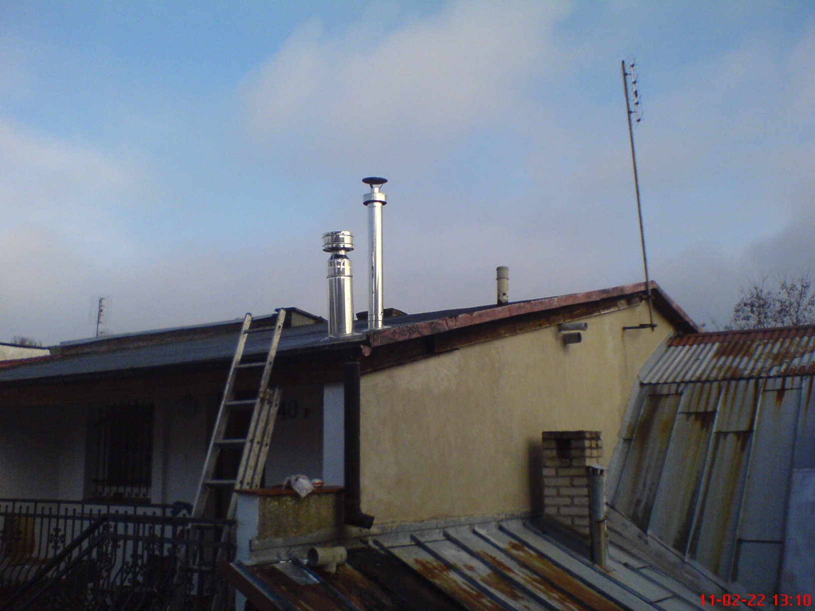 Dwa nowe, błyszczące, metalowe kominy wentylacyjne wystające ponad dach budynku z drabina opartą o ścianę. W tle błękitne niebo z chmurami.