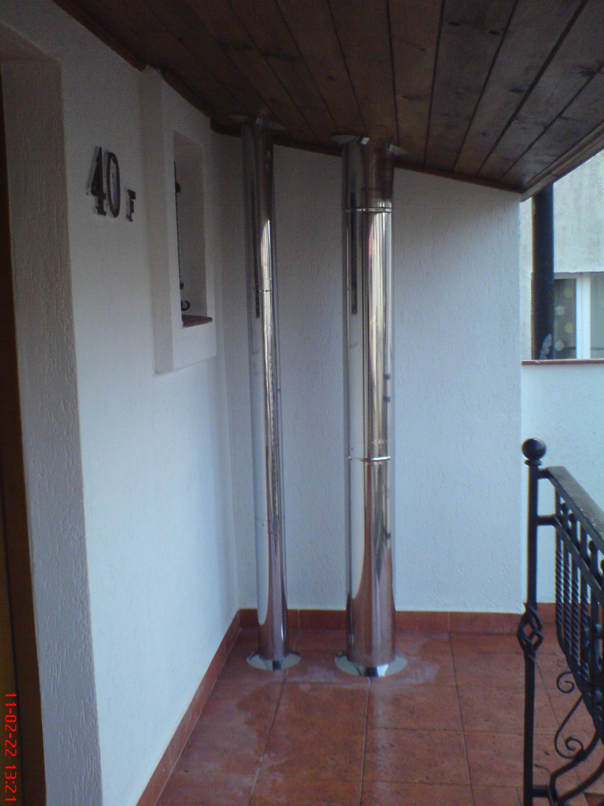 Dwa chromowane kominy odprowadzające spaliny, wystające z podłogi balkonu z brązowych płytek, z drewnianym sufitem i fragmentem metalowej balustrady.