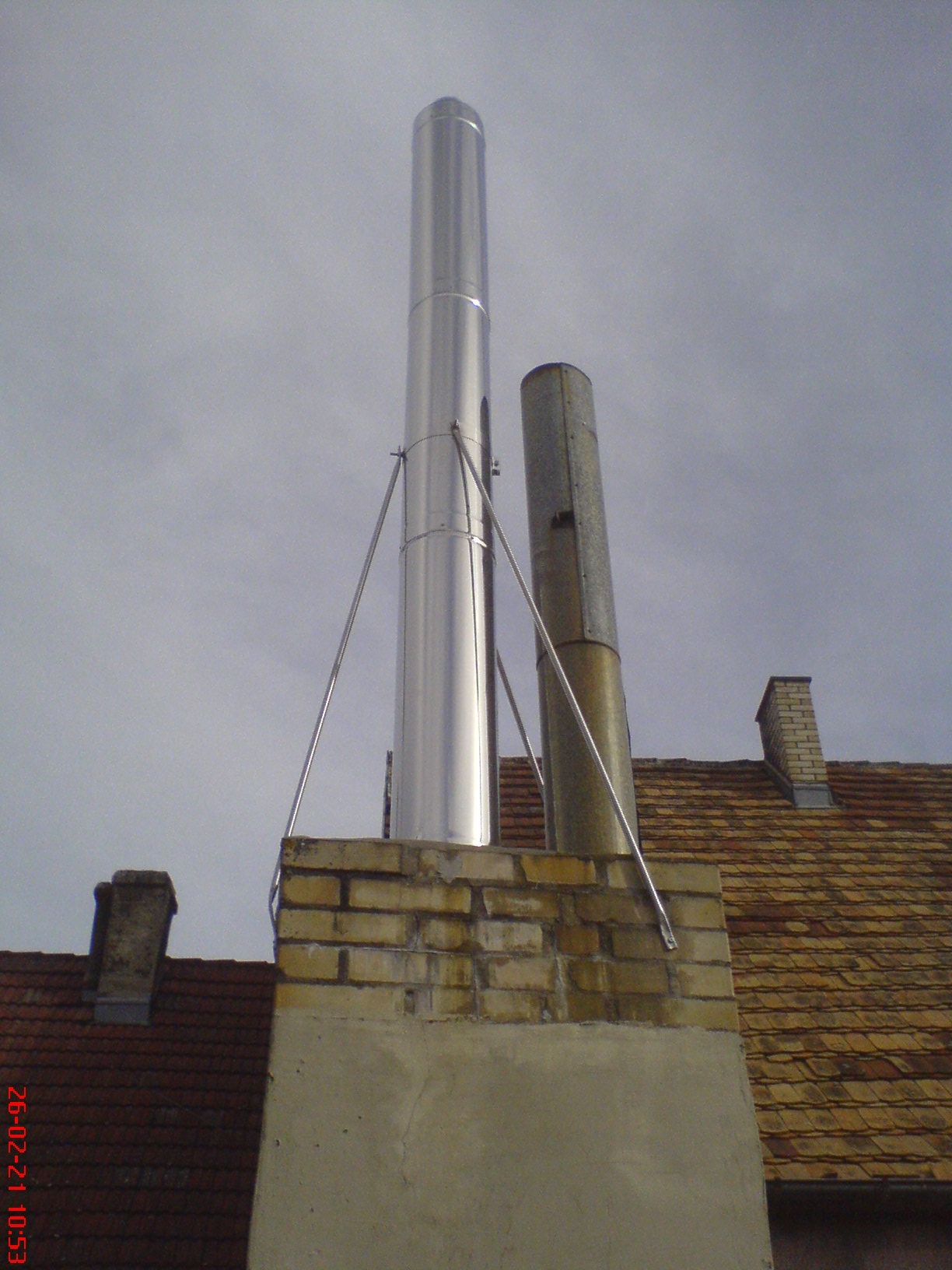 Nowy, błyszczący komin ze stali nierdzewnej zamontowany obok starego, skorodowanego komina na ceglanym dachu z widocznymi mocowaniami.