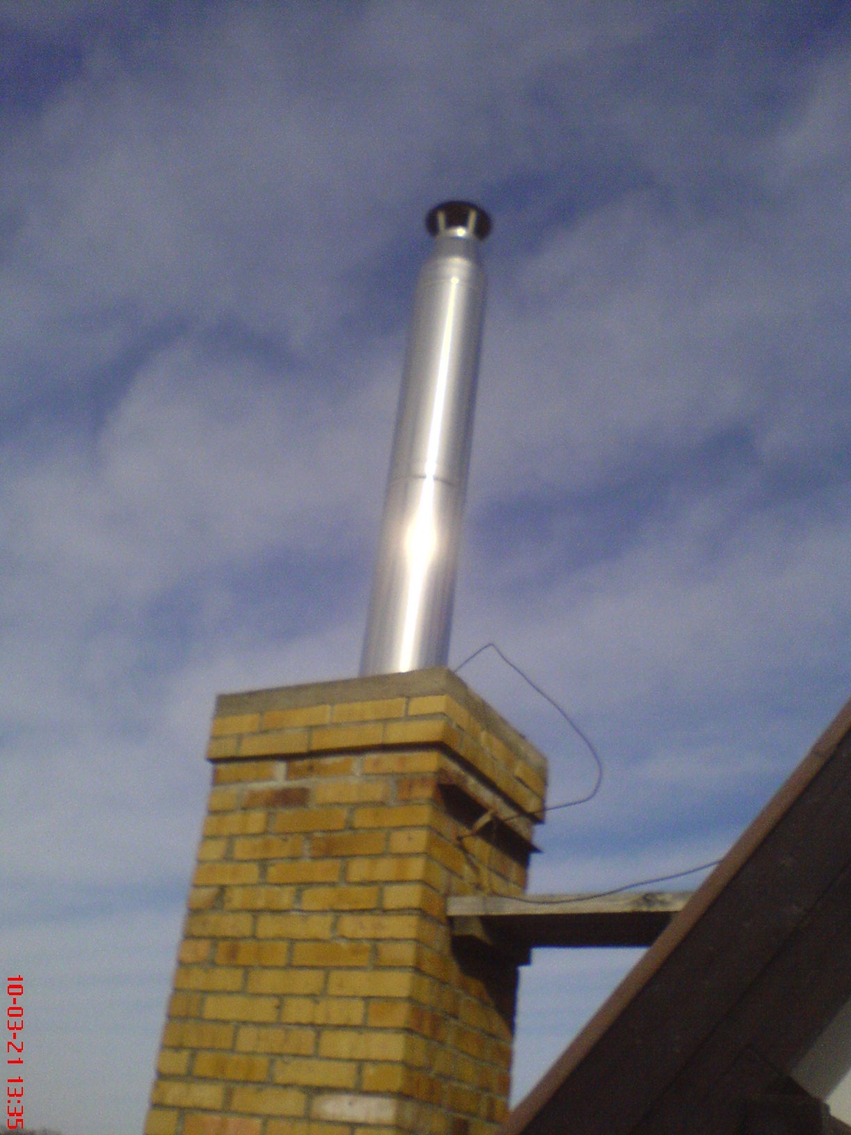 Komin z cegły z metalowym wkładem kominowym, widoczny fragment dachu, na tle błękitnego nieba z chmurami.