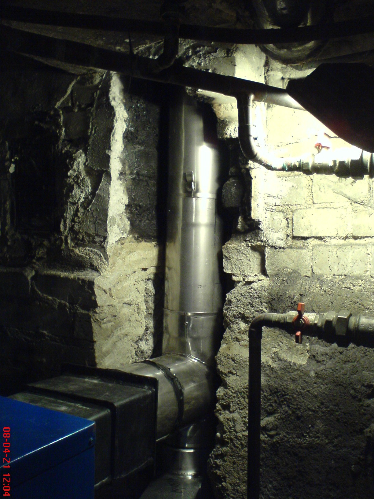 Metalowy przewód kominowy w piwnicy z surowymi, kamiennymi ścianami, widoczne rury instalacyjne i fragment niebieskiej obudowy urządzenia.