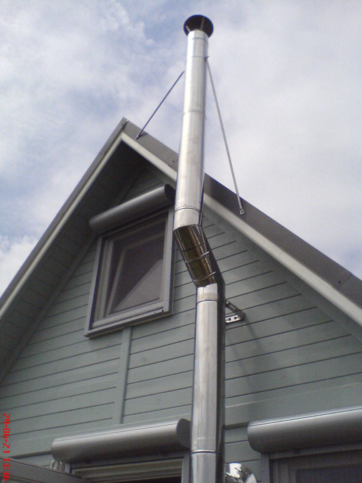 Zewnętrzny, stalowy system kominowy, zamocowany do bocznej ściany domu z drewnianą elewacją i oknem na poddaszu.