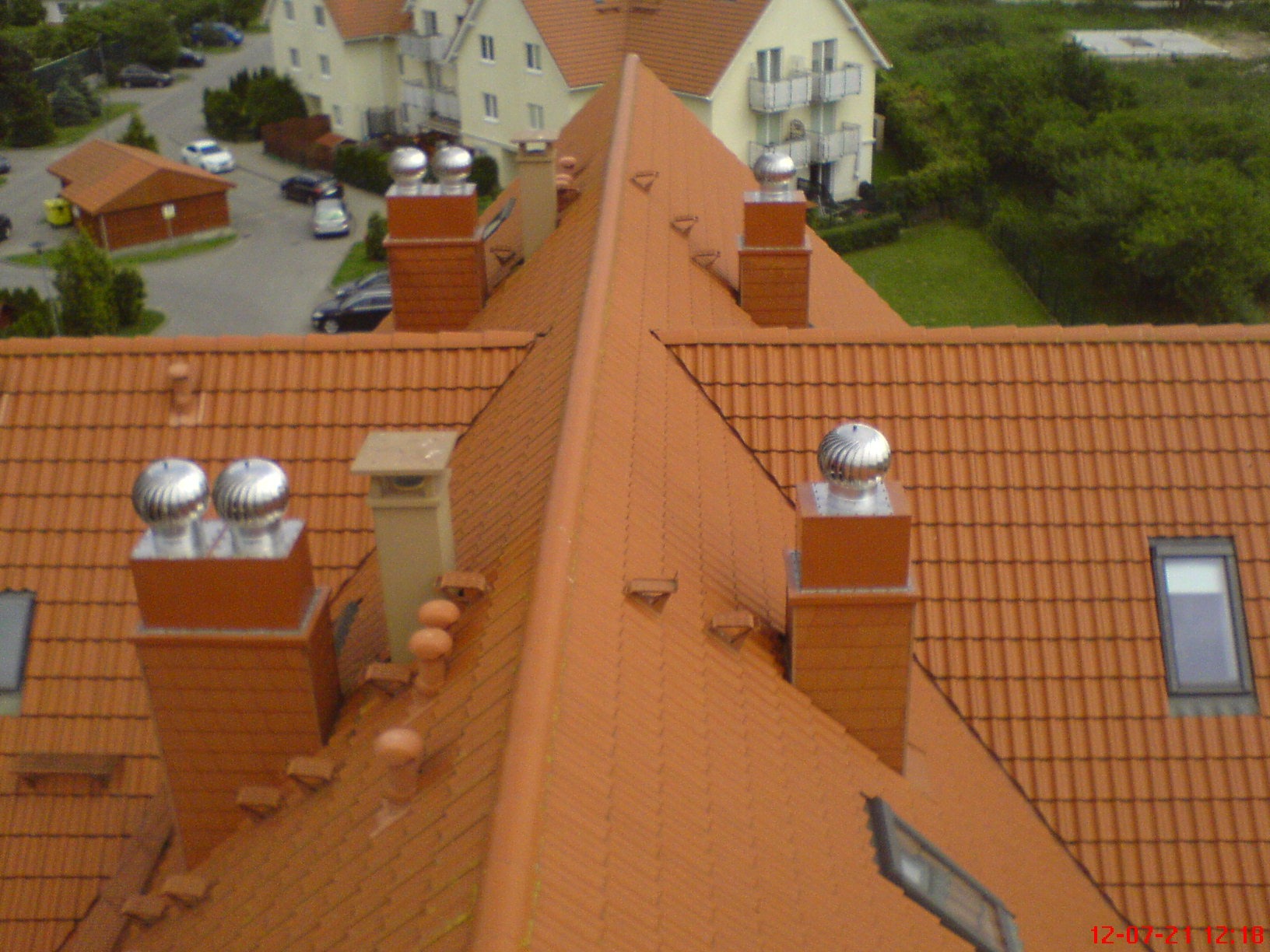 Czerwony dach wielospadowy z kominami, wentylatorami turbinowymi i oknami dachowymi, widok z góry na tle zabudowy mieszkalnej i zieleni.