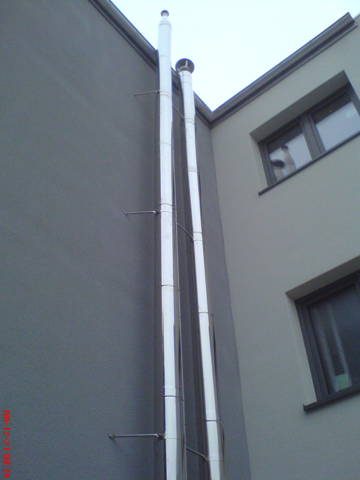 Dwa błyszczące, metalowe kominy zamontowane na zewnętrznej ścianie budynku, z widocznymi wspornikami mocującymi i elementami wykończeniowymi na szczycie.