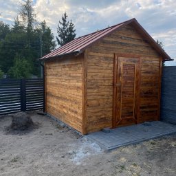 Styl Dach Mariia Popovych - Nowo postawiona drewniana szopa z brązowym dachem z blachy trapezowej, widoczne obróbki blacharskie, kostka brukowa przed wejściem i sterta ziemi obok.