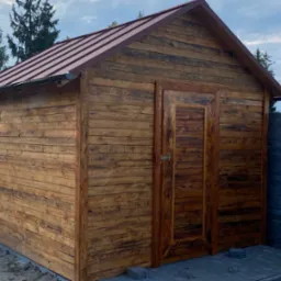 Drewniany domek narzędziowy z brązowym metalowym wykończeniem dachu i rynnami, widok z przodu, na tle częściowo zachmurzonego nieba i ogrodzenia.