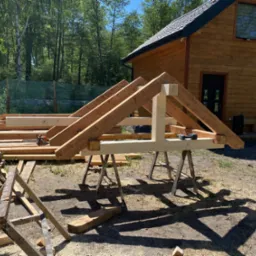 Konstrukcja drewnianej więźby dachowej na kozłach montażowych przed domem z bali, w tle stos desek i narzędzia.