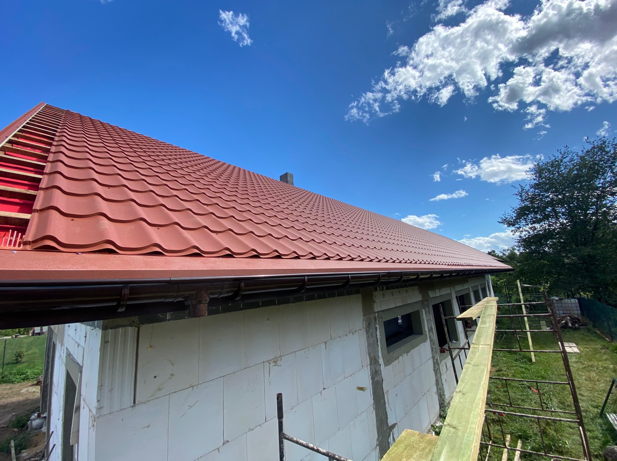 Czerwona dachówka ceramiczna na budynku w trakcie budowy, widok z dołu na tle błękitnego nieba z chmurami, widoczne rynny i elementy konstrukcji dachu.