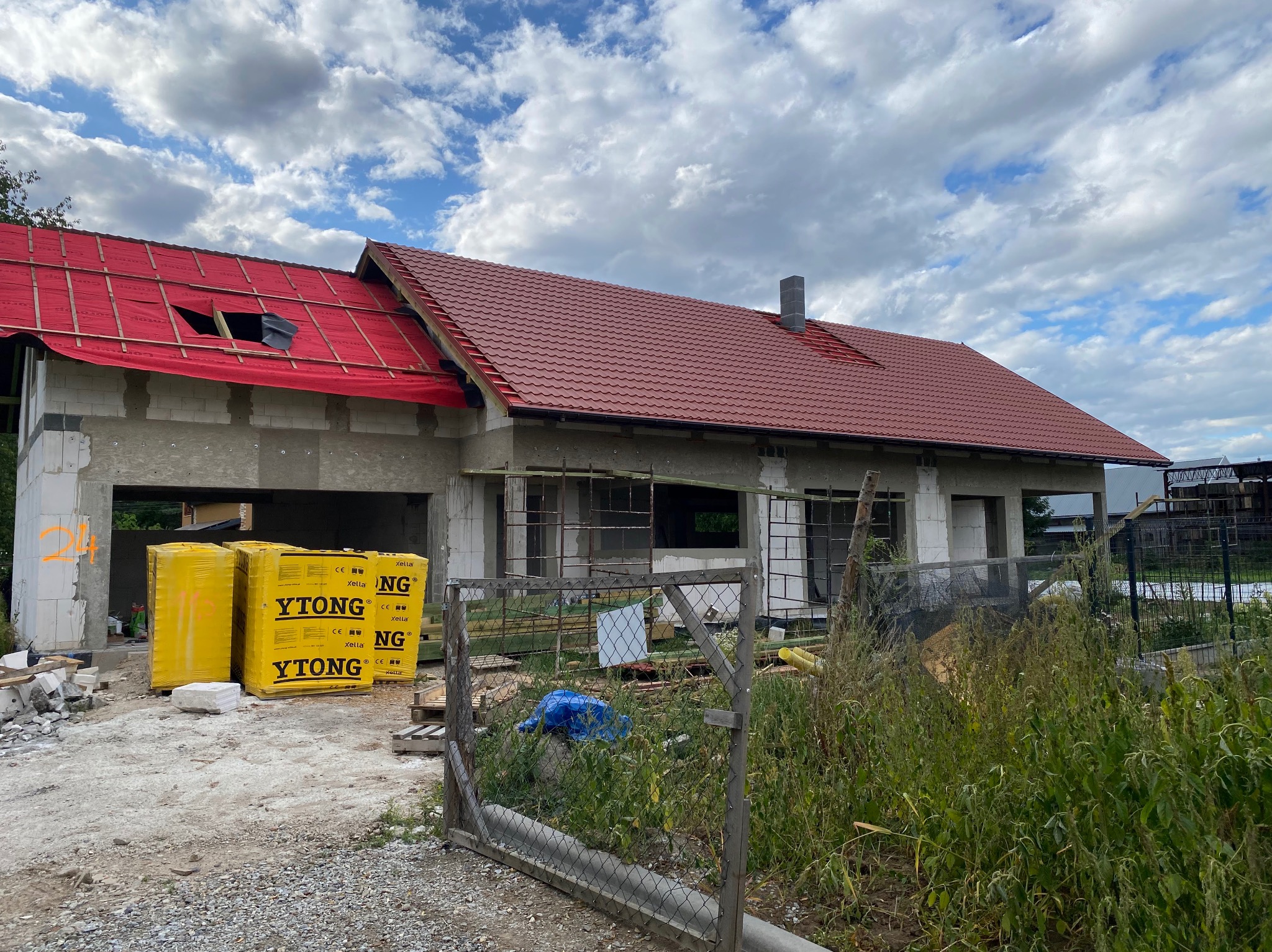 Budowa domu w trakcie krycia dachu czerwoną dachówką, widoczne materiały budowlane YTONG i membrana dachowa, częściowo odkryta więźba dachowa.