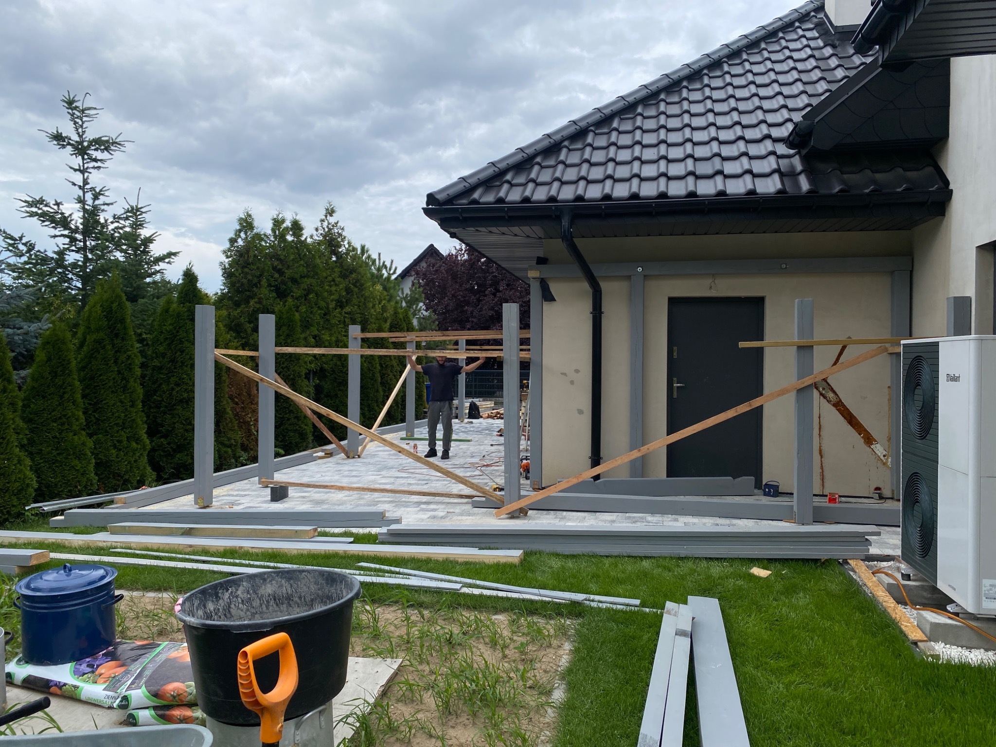 Montaż drewnianej konstrukcji z szarymi słupami na tle domu z ciemnym dachem, widoczna osoba przy pracy, przygotowane elementy konstrukcyjne leżą na trawie i kostce brukowej.