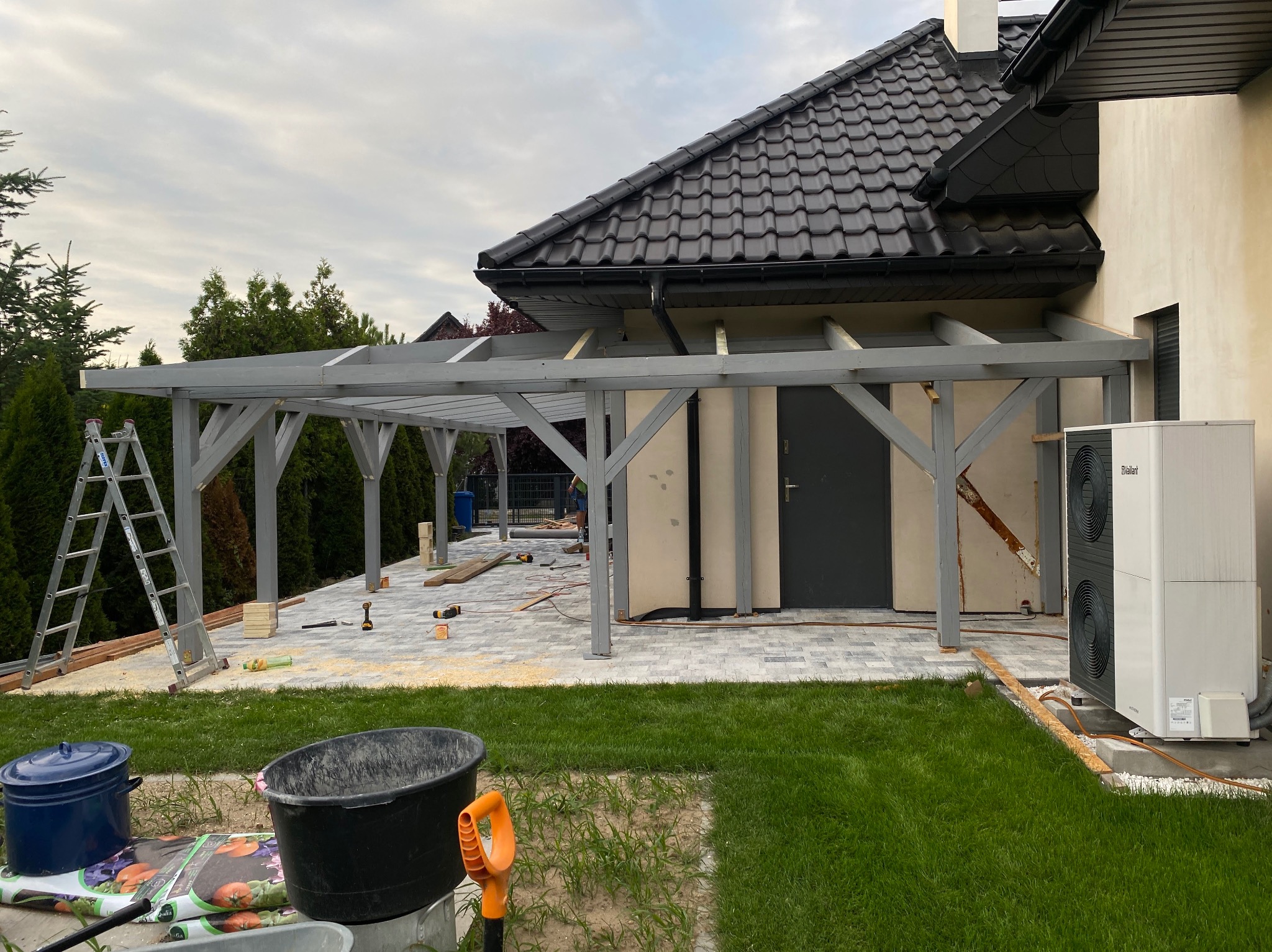 Szara, drewniana konstrukcja zadaszenia tarasu w trakcie budowy, widoczna drabina, narzędzia i materiały budowlane na kostce brukowej, dom z ciemną dachówką w tle, obok pompa ciepła.