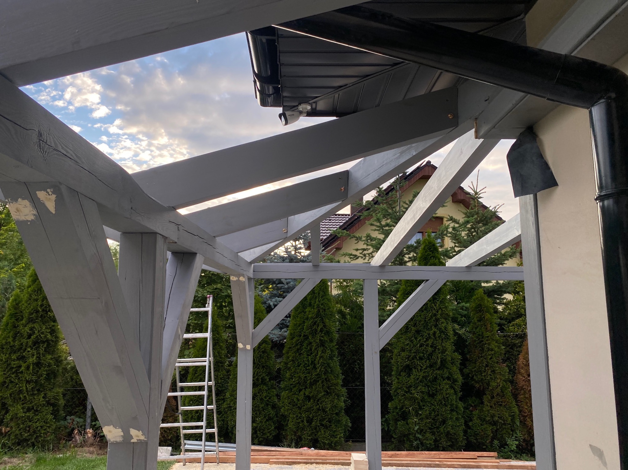 Surowa, szara konstrukcja drewniana z widoczną więźbą, fragmentem orynnowania i drabiną opartą o jeden ze słupów, na tle zielonych drzew i błękitnego nieba z chmurami.