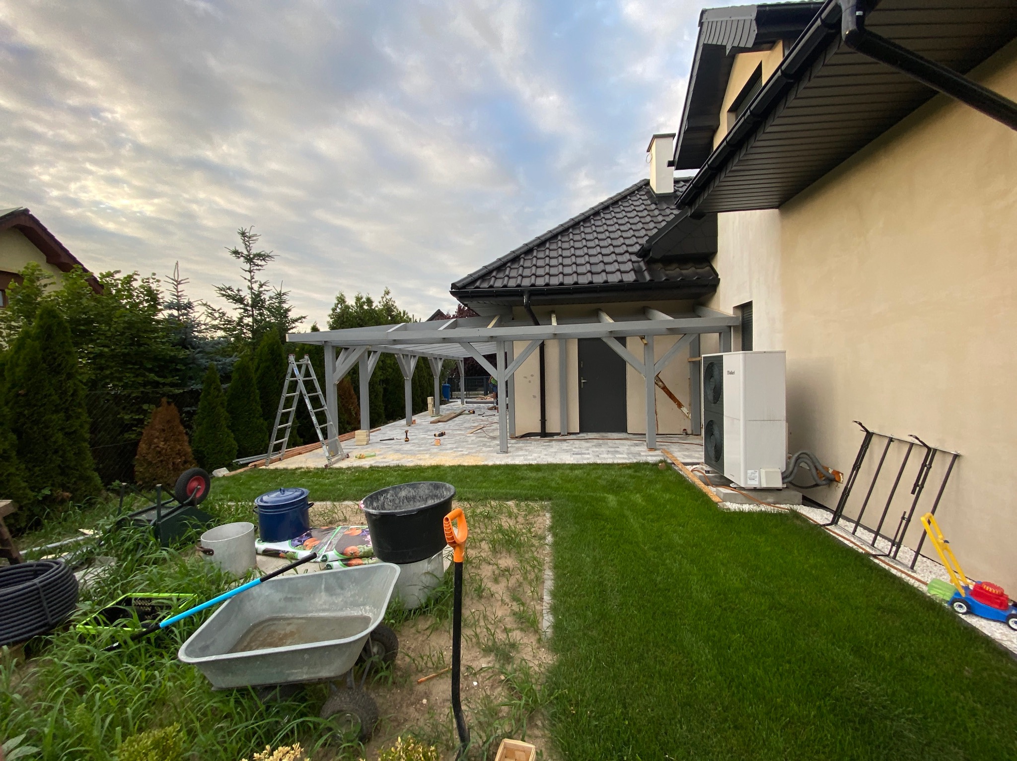 Montaż drewnianej pergoli przy domu z ciemnym dachem, widoczne narzędzia i materiały budowlane na trawniku, drabina oparta o konstrukcję, w tle zielony ogród i pochmurne niebo.