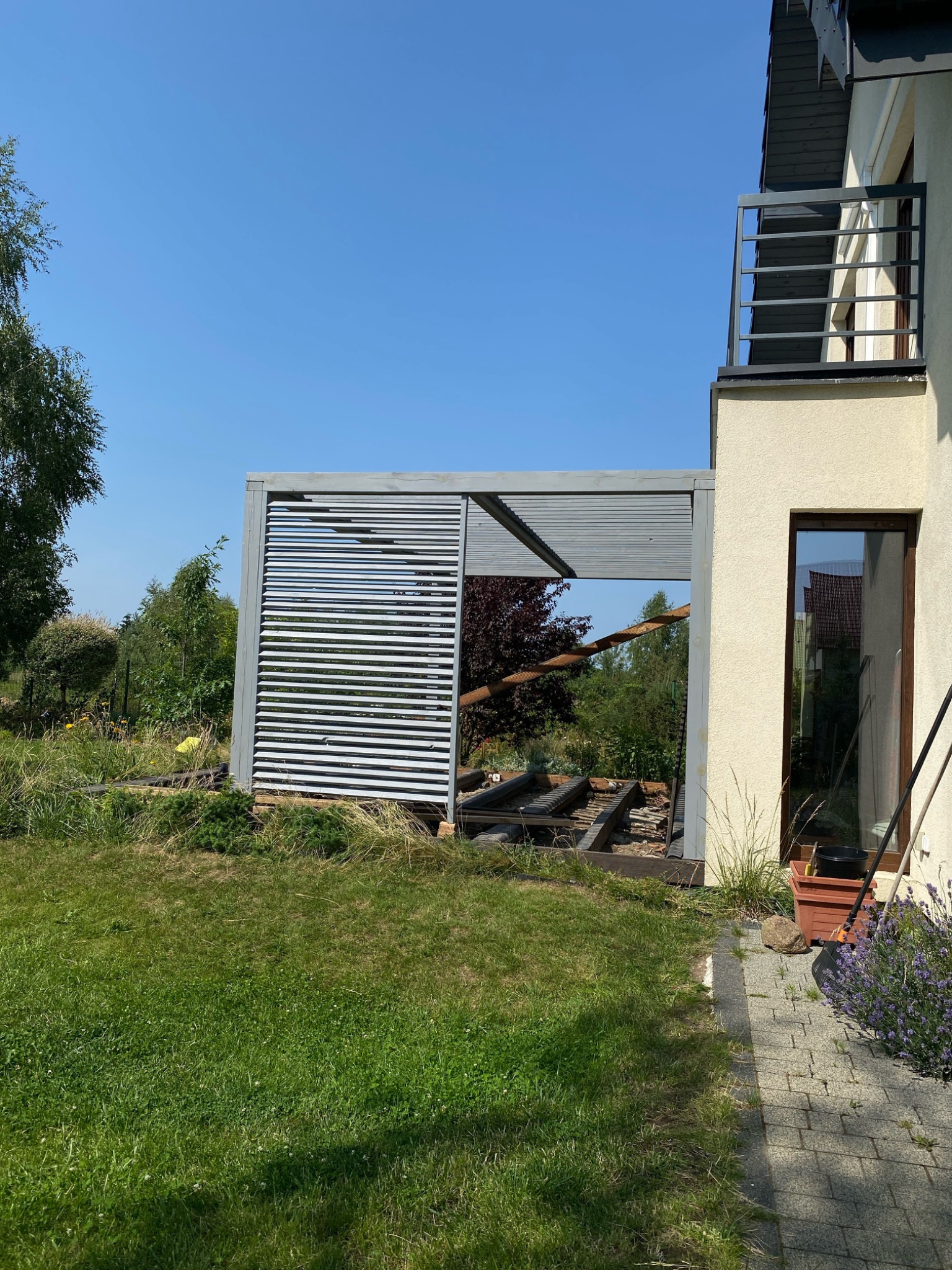 Szara pergola lamelowa w trakcie montażu, widoczna konstrukcja dachu, na trawniku leżą narzędzia, obok domu z balkonem.
