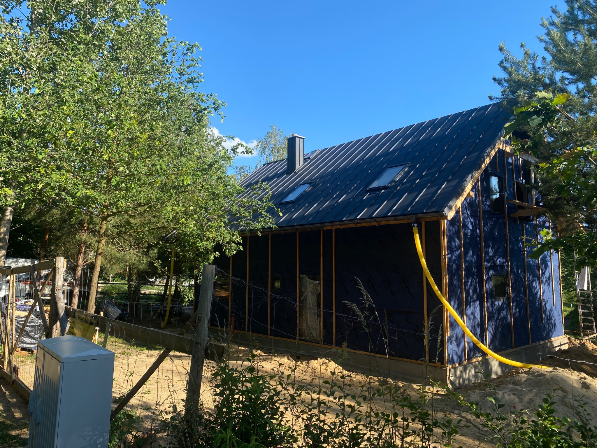 Nowoczesny dom w trakcie budowy, z czarnym, metalowym pokryciem i oknami dachowymi, widoczna konstrukcja ścian i żółty przewód doprowadzony do budynku.