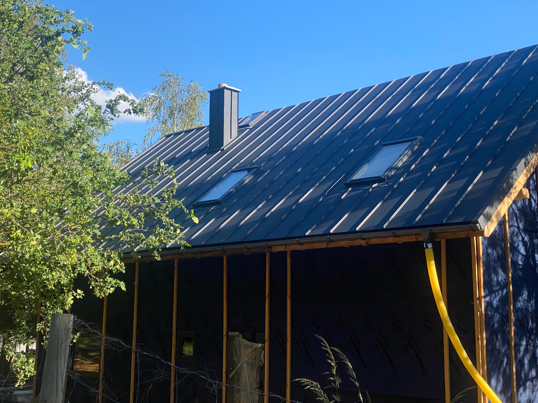 Nowoczesny dom z czarnym dachem z blachy na rąbek stojący, dwa okna dachowe, komin i żółta rura spustowa, widok z dołu na tle błękitnego nieba.