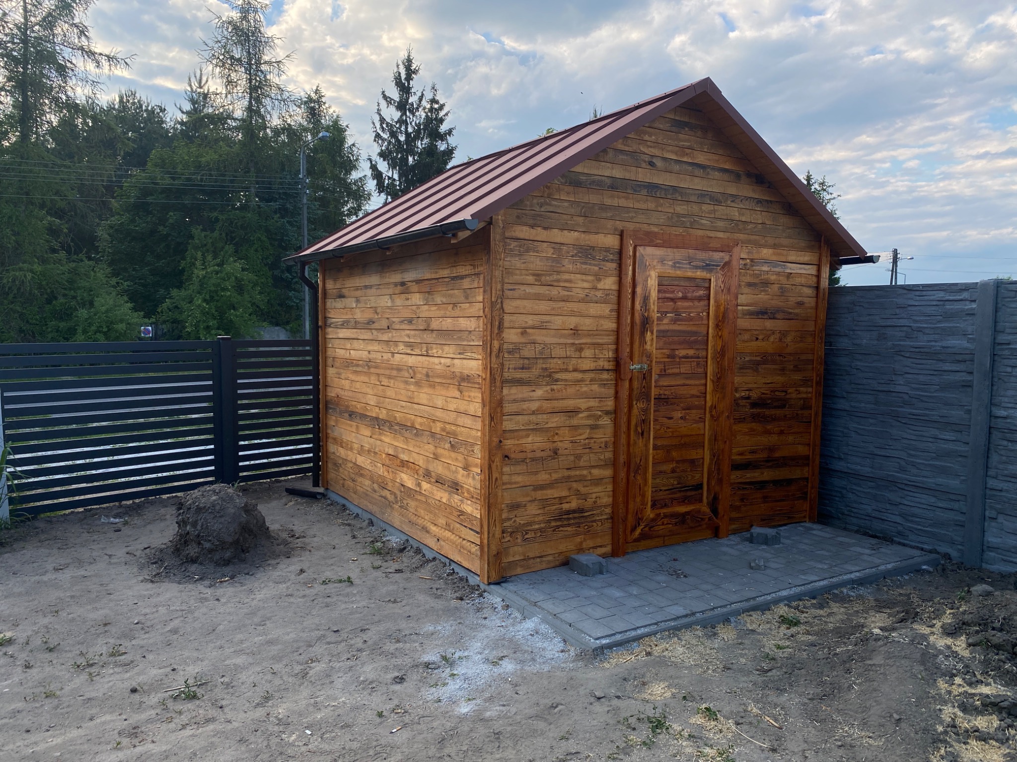 Nowo postawiona drewniana szopa z brązowym dachem z blachy trapezowej, widoczne obróbki blacharskie, kostka brukowa przed wejściem i sterta ziemi obok.