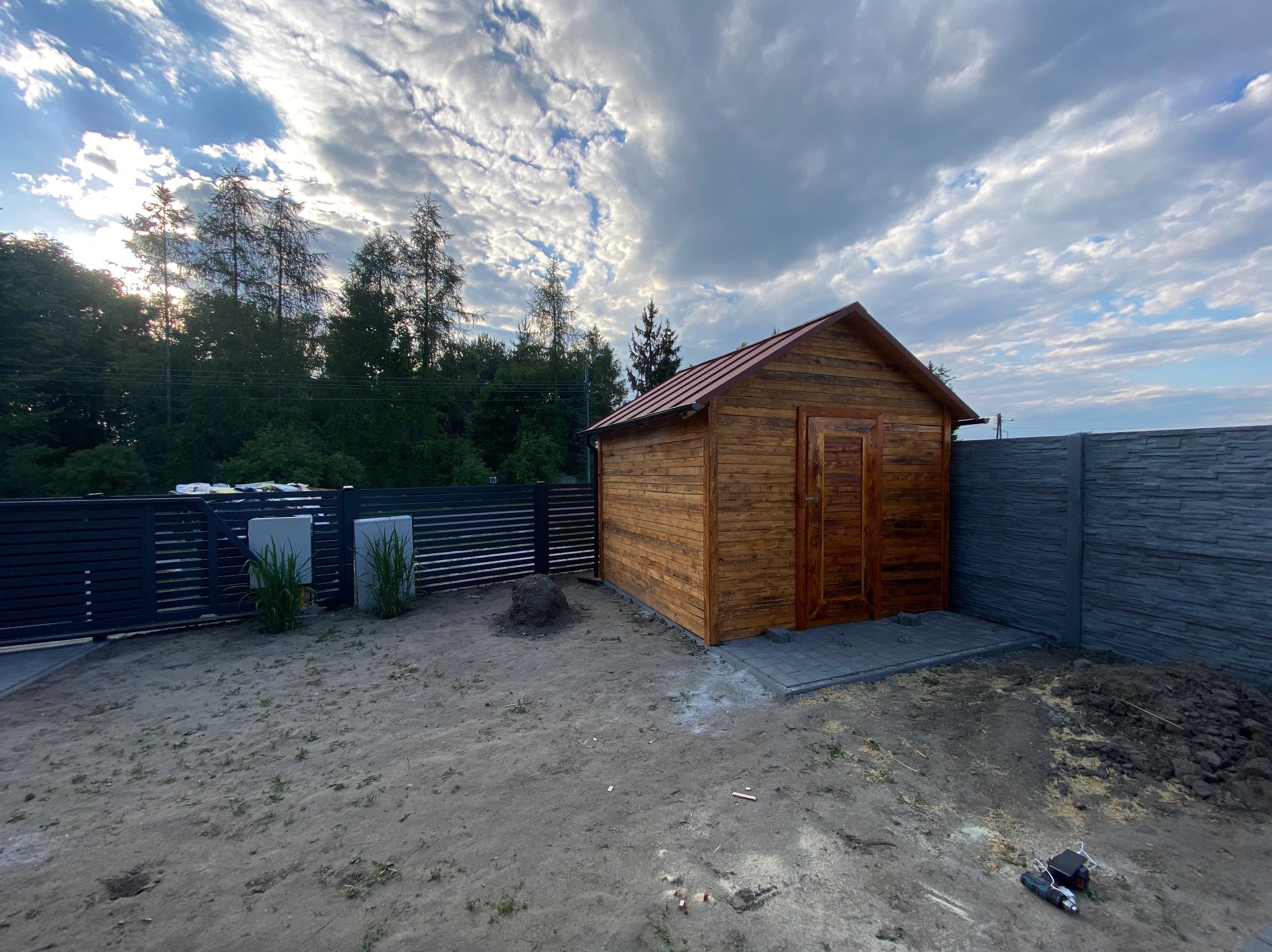 Nowo postawiony, drewniany budynek gospodarczy z czerwoną blachą na dachu, widoczny na tle pochmurnego nieba i ogrodzenia.