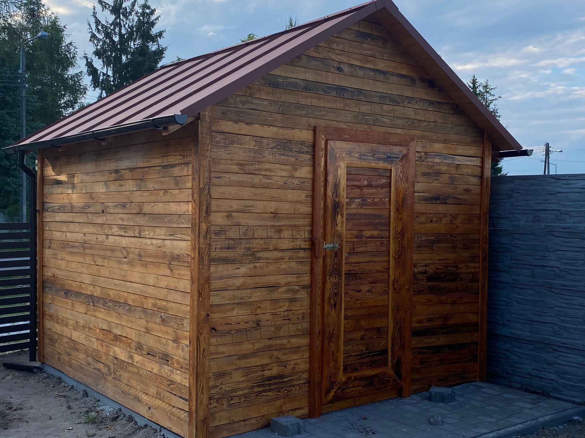 Drewniany domek narzędziowy z brązowym metalowym wykończeniem dachu i rynnami, widok z przodu, na tle częściowo zachmurzonego nieba i ogrodzenia.