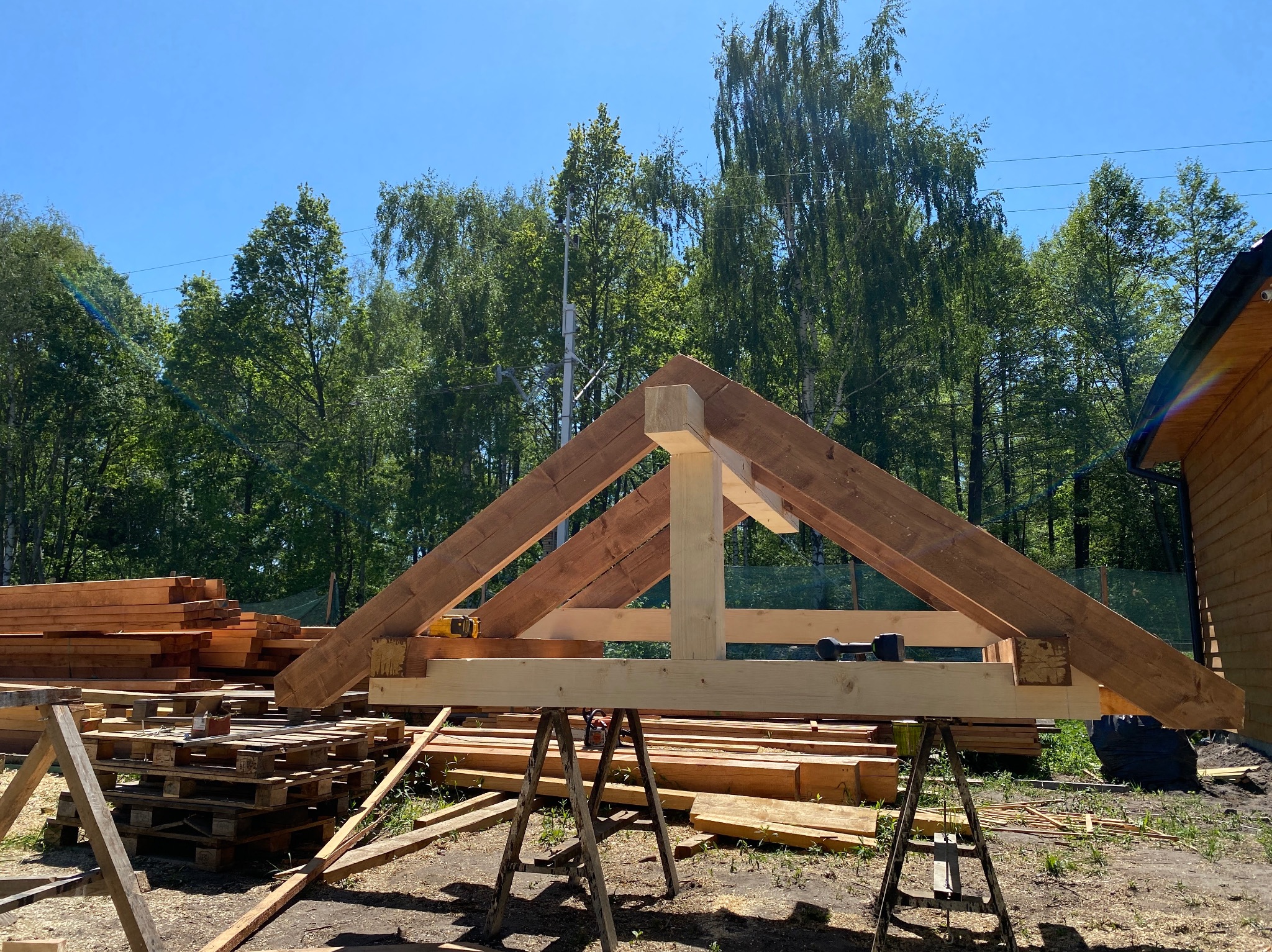 Wstępny montaż drewnianej więźby dachowej na metalowych kozłach, w tle stos drewna, palety i zieleń drzew.