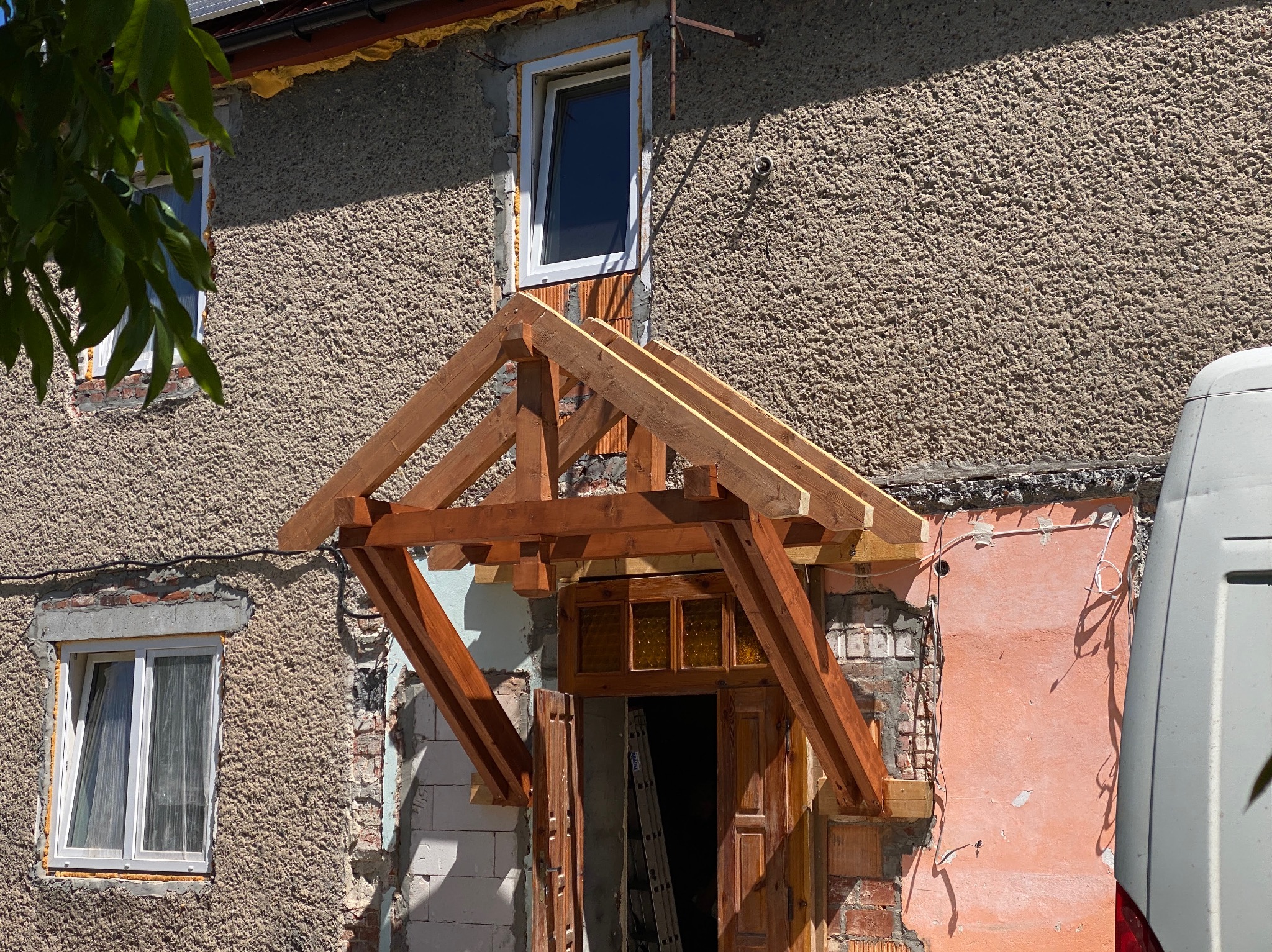 Drewniana konstrukcja zadaszenia nad wejściem do domu w trakcie montażu, widoczne surowe drewno i elementy mocujące, ściany z uszkodzonym tynkiem i widocznymi cegłami, drabina oparta o futrynę drzwi.