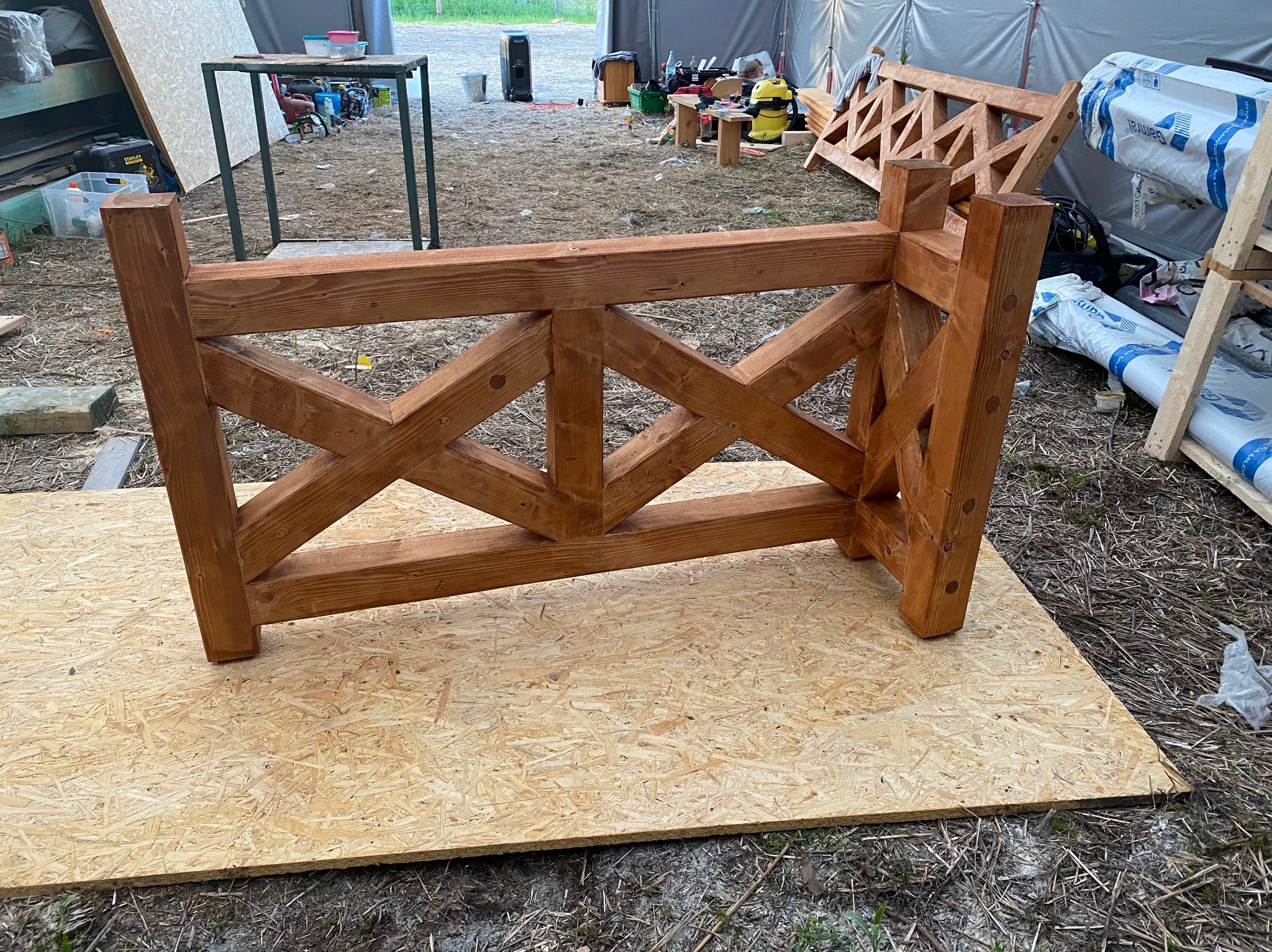 Fragment drewnianej konstrukcji z krzyżującymi się belkami, prawdopodobnie elementu więźby dachowej, leżący na płycie OSB w warunkach budowy, z widocznymi narzędziami i materiałami w tle.