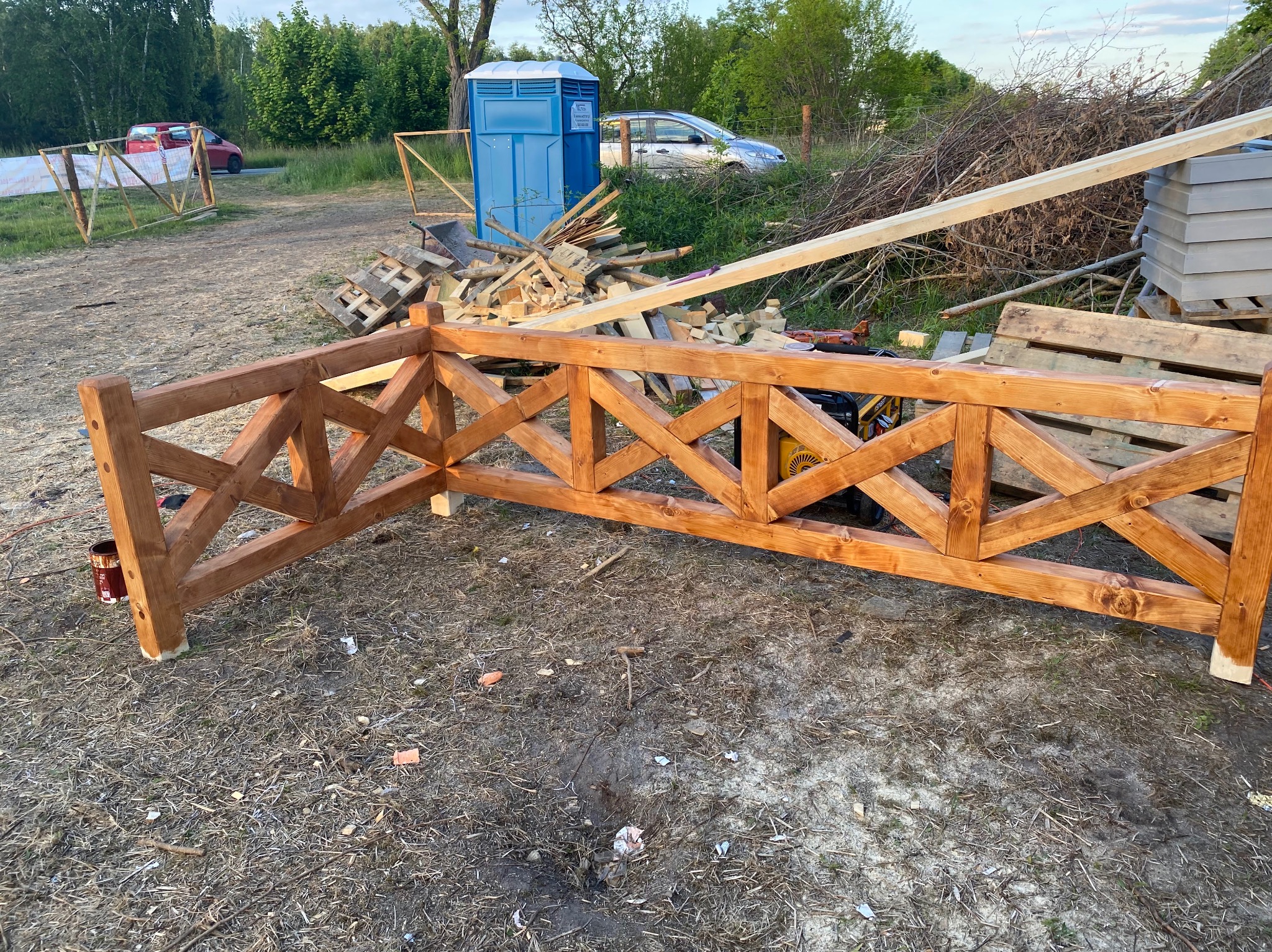 Fragment drewnianej konstrukcji ogrodzenia w stylu rustykalnym, z widocznym słoikiem farby i przenośną toaletą w tle, na placu budowy.