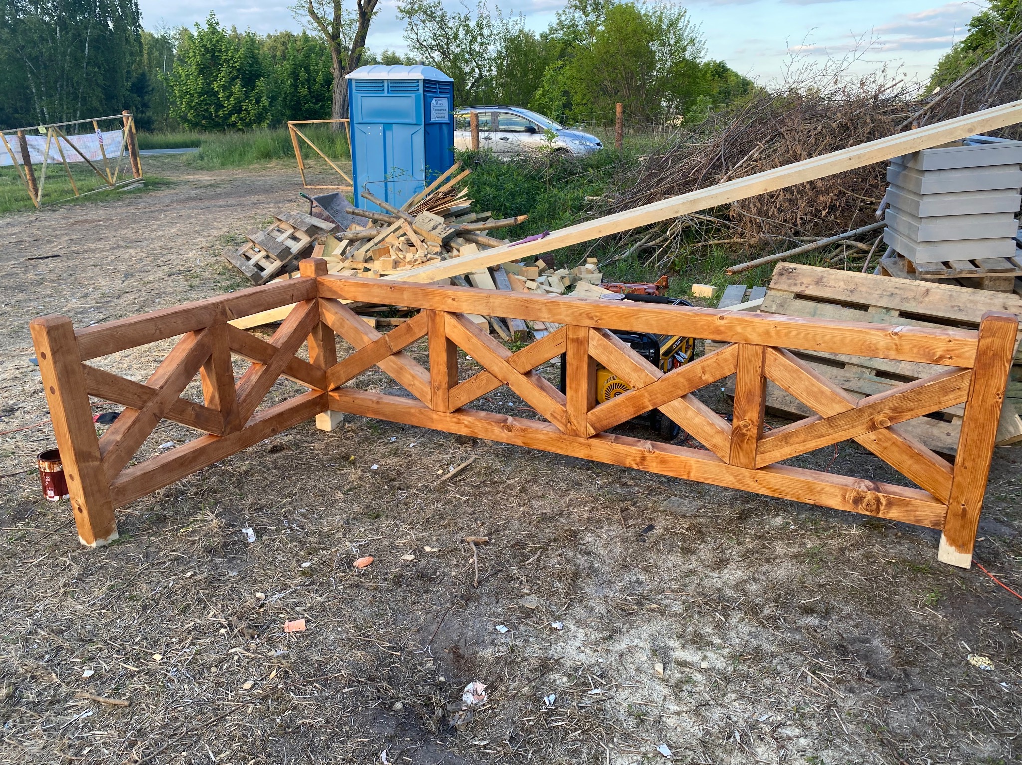 Fragment nowej, drewnianej balustrady z krzyżowym wzorem, świeżo pomalowanej bejcą, w tle przenośna toaleta i materiały budowlane na placu budowy.