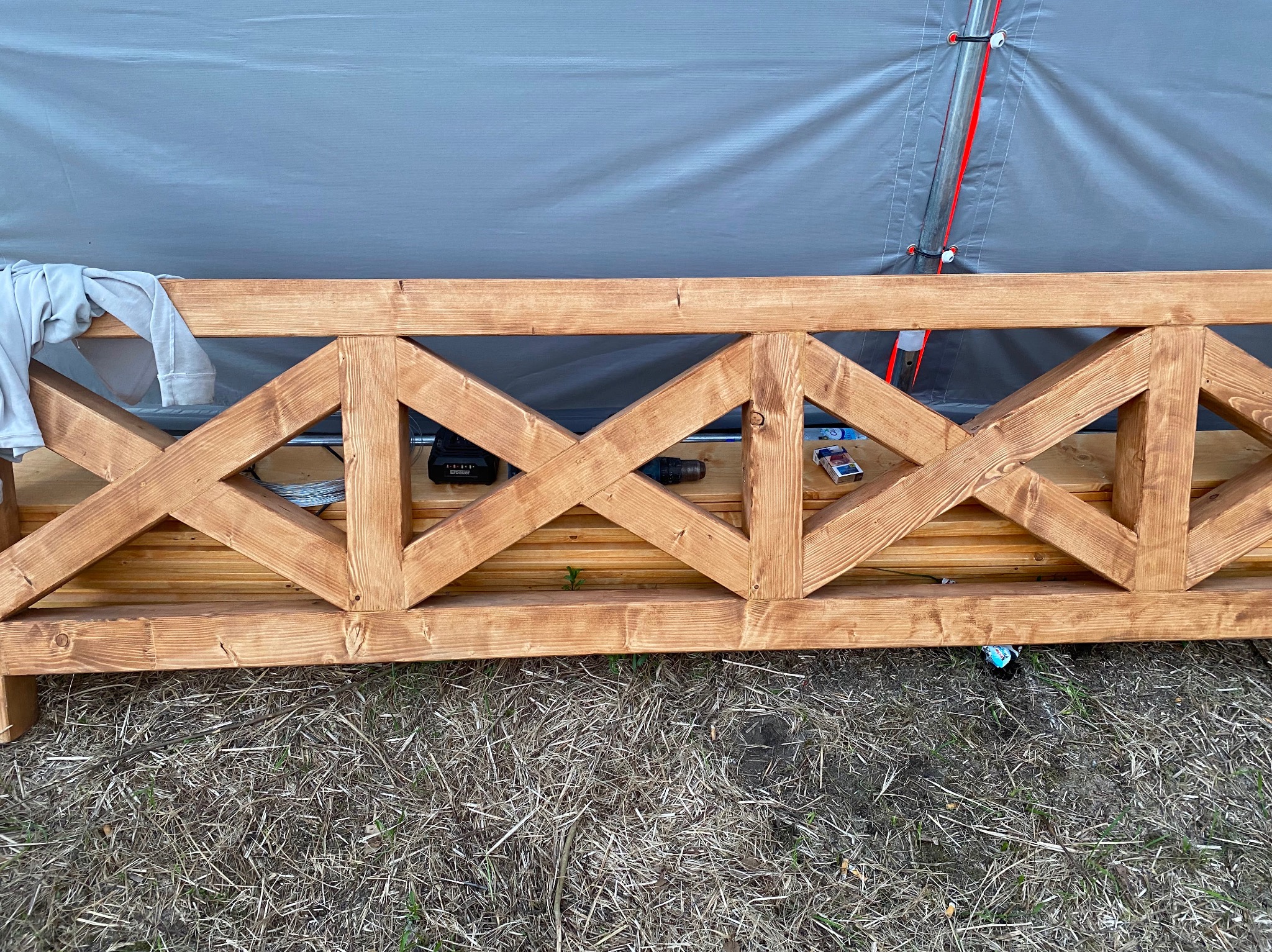 Fragment drewnianej konstrukcji z ozdobnymi krzyżulcami, widoczny akumulatorowy wkrętak, pudełko zapałek, ubranie i butelka pod spodem, na tle szarej plandeki.