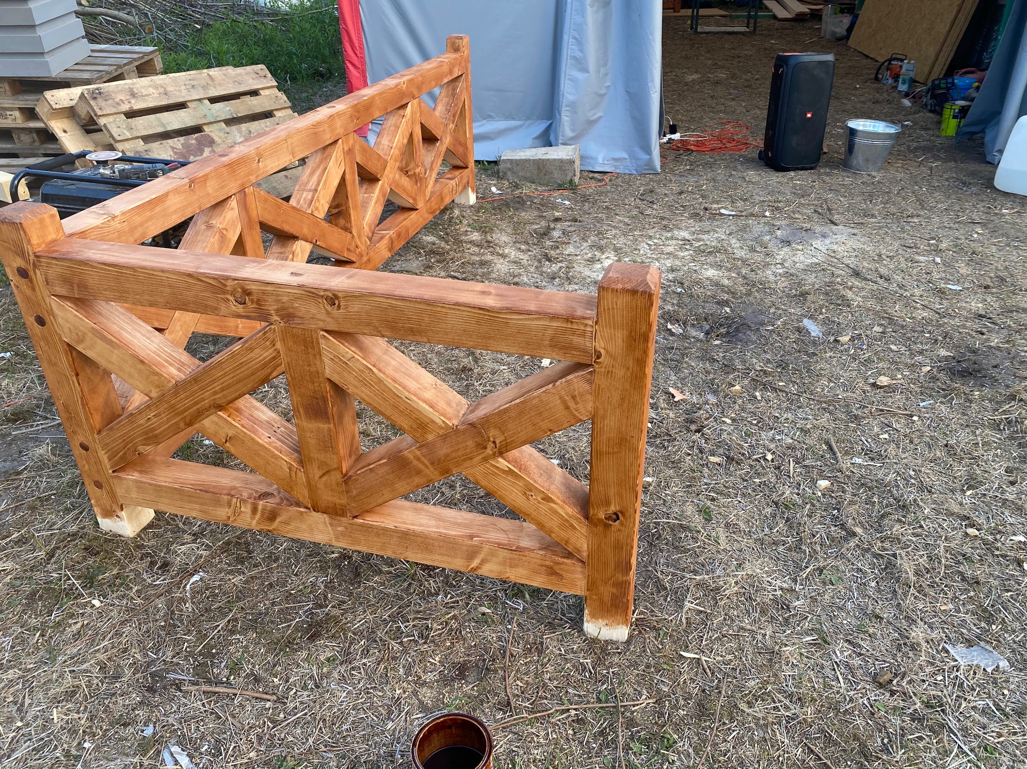 Fragment drewnianej konstrukcji kratownicowej, prawdopodobnie elementu więźby dachowej, świeżo zaimpregnowany brązowym preparatem, widoczny pędzel i puszka z impregnatem, w tle palety i sprzęt...