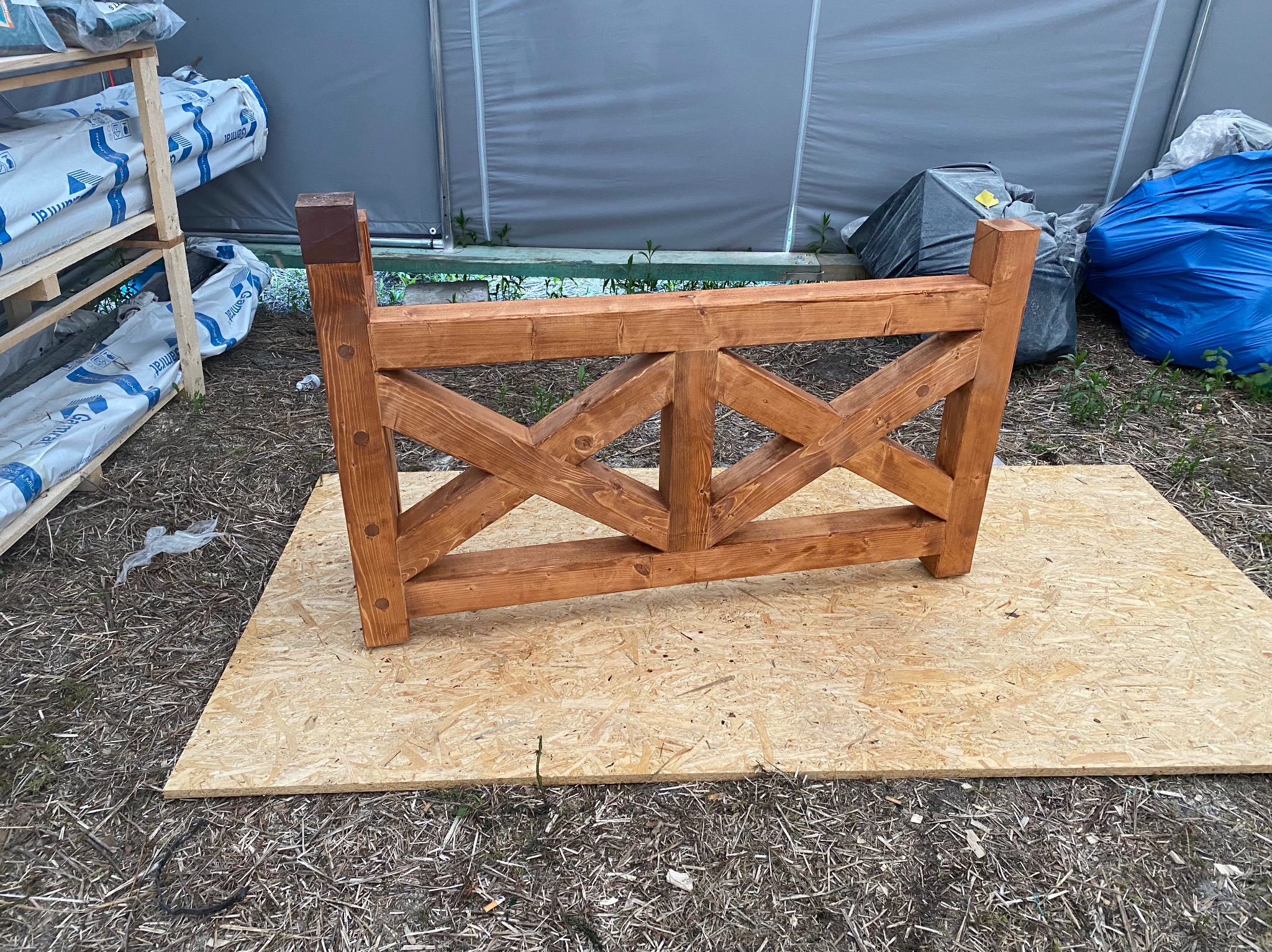 Drewniana konstrukcja kratowa, prawdopodobnie element więźby dachowej, leży na płycie OSB, w tle materiały budowlane i zarośla.