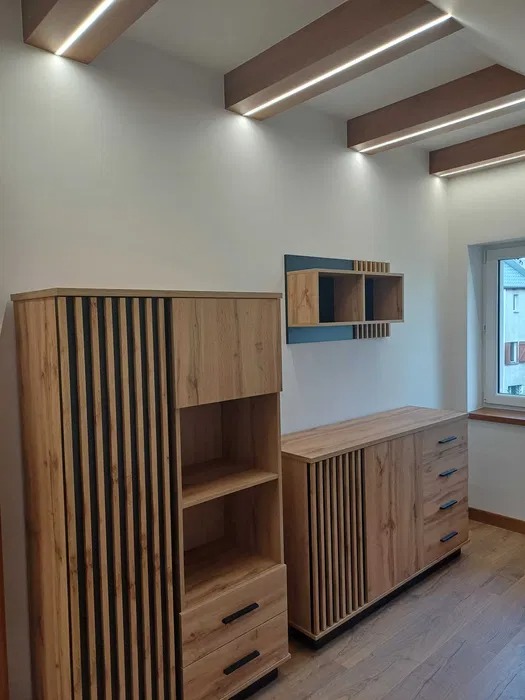 Nowoczesne meble z drewnianymi lamelami w jasnym wnętrzu z podświetlanym sufitem, widok na regał, komodę i półki wiszące.