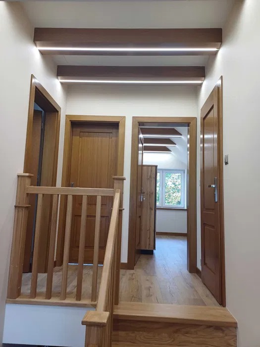 Wnętrze domu z drewnianymi schodami, balustradą i trzema parami drewnianych drzwi, oświetlone listwami LED pod sufitem.