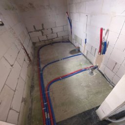 ZUH Broser - Instalacja wodna i grzewcza w surowym pomieszczeniu z białych bloczków. Czerwone i niebieskie rury prowadzone po podłodze i ścianach. Widoczne elementy instalacji kanalizacyjnej.
