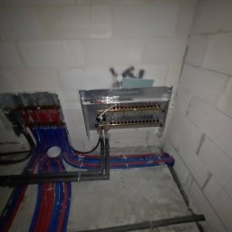 ZUH Broser - Rozdzielacz ogrzewania podłogowego zamontowany na ścianie z bloczków, z widocznymi rurami w kolorach niebieskim i czerwonym. Instalacja w trakcie montażu, surowy stan.