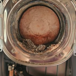 ZUH Broser - Zanieczyszczony palnik pieca gazowego z widocznymi osadami i korozją. Miedziane rurki instalacji hydraulicznej w dolnej części kadru. Wnętrze pieca w zbliżeniu.