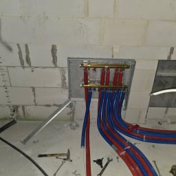 ZUH Broser - Rozdzielacz ogrzewania podłogowego z rurami PEX w kolorze czerwonym i niebieskim, zamontowany na ścianie z betonu komórkowego. Widoczne narzędzia hydrauliczne.
