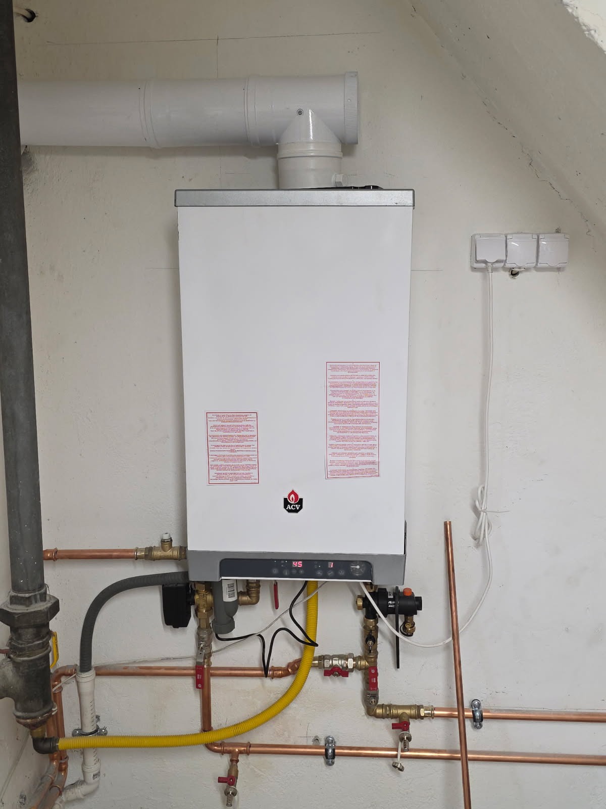 Biały piec gazowy ACV zamontowany na ścianie z widocznymi rurami miedzianymi i żółtym przewodem gazowym. Nad piecem biały komin, obok gniazdka elektryczne.