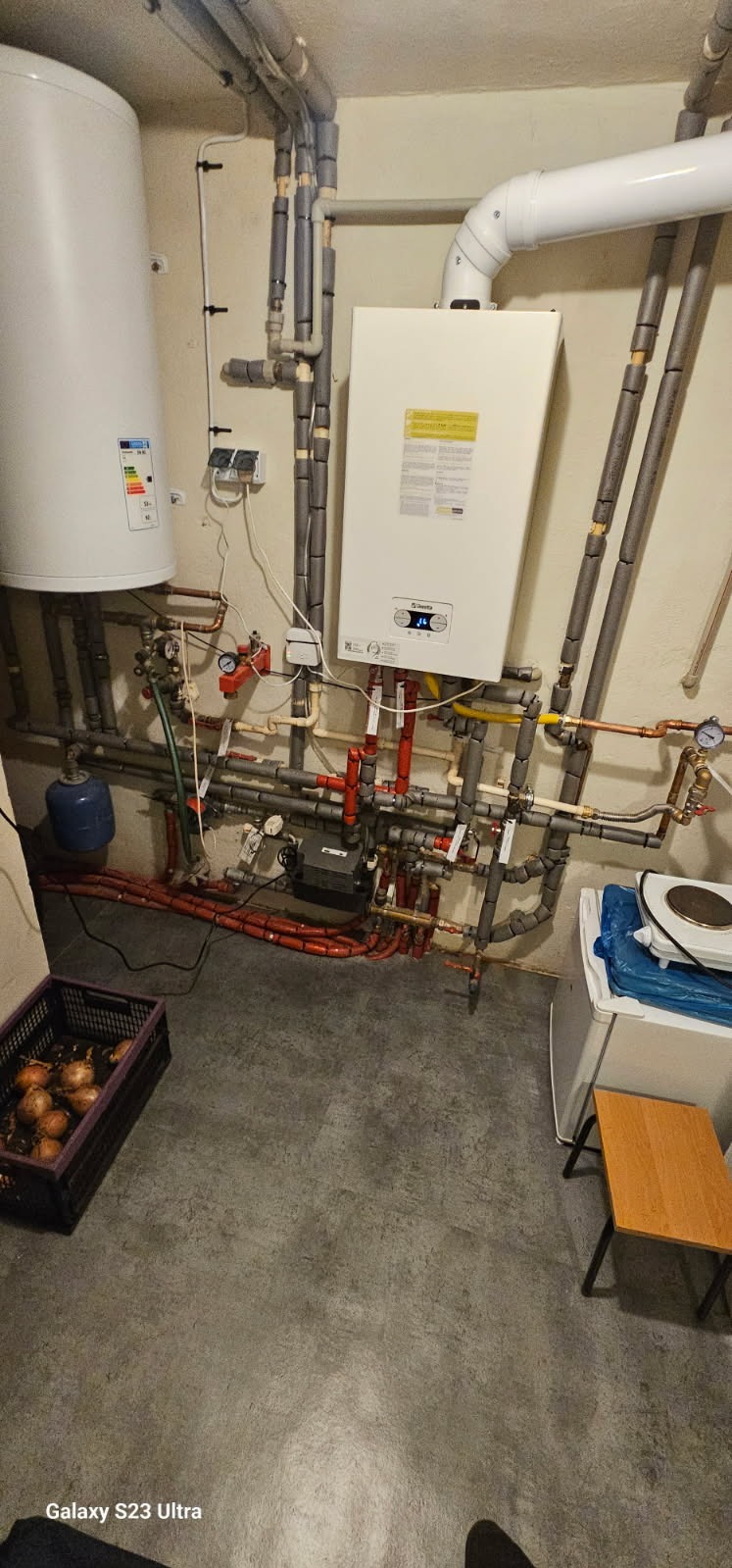 Złożona instalacja CO z piecem gazowym Beretta, zbiornikiem i licznymi rurami w piwnicy. Widoczne liczniki, zawory i izolacje. Na podłodze skrzynka z cebulą.