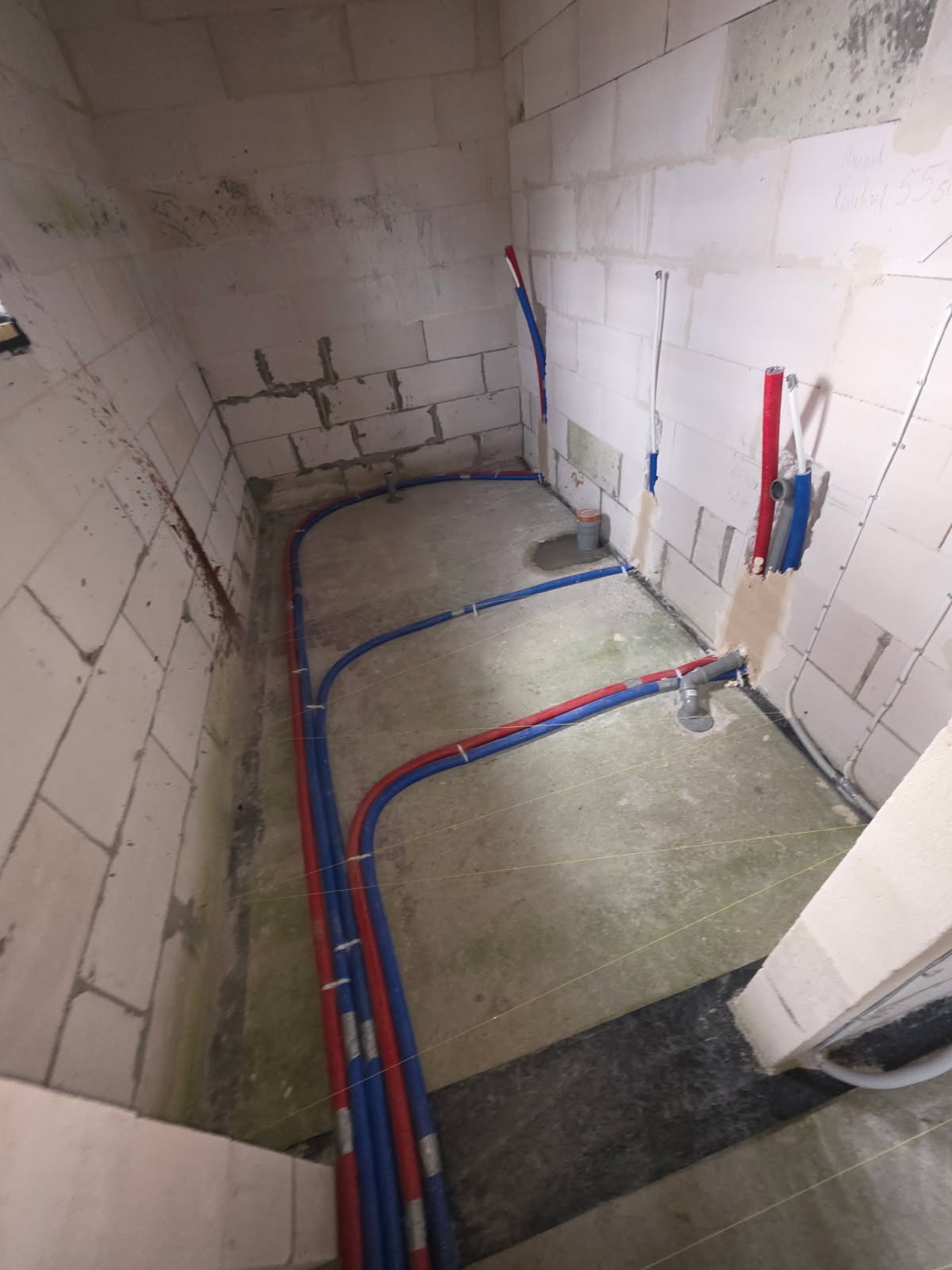 Instalacja wodna i grzewcza w surowym pomieszczeniu z białych bloczków. Czerwone i niebieskie rury prowadzone po podłodze i ścianach. Widoczne elementy instalacji kanalizacyjnej.