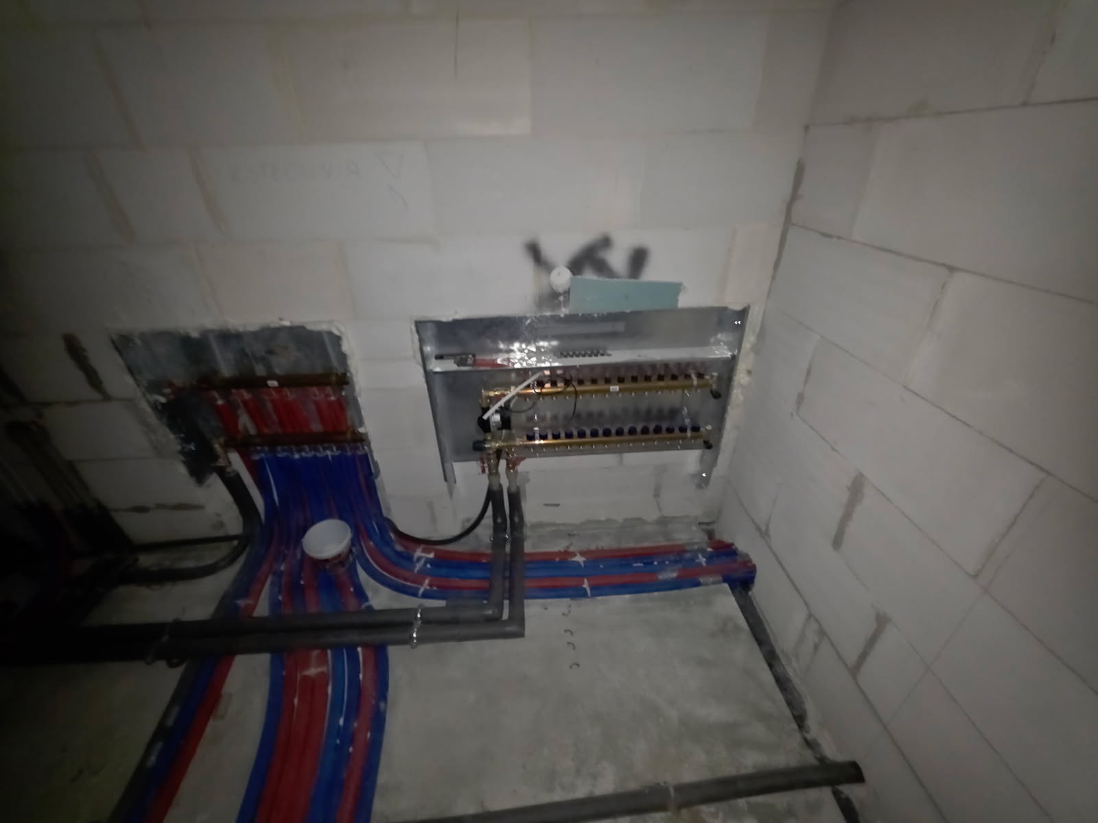 Rozdzielacz ogrzewania podłogowego zamontowany na ścianie z bloczków, z widocznymi rurami w kolorach niebieskim i czerwonym. Instalacja w trakcie montażu, surowy stan.