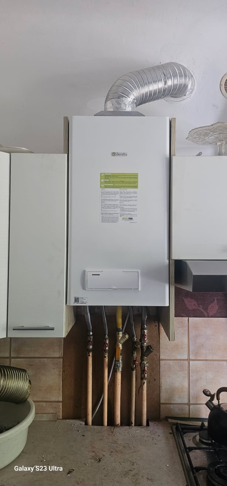 Biały piec gazowy Beretta zamontowany między szafkami kuchennymi, widoczne rury doprowadzające gaz i wodę, oraz aluminiowy przewód spalinowy.