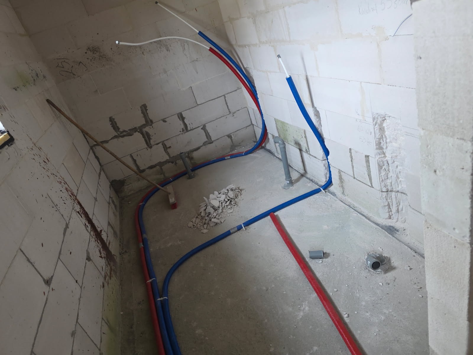 Instalacja wodna i kanalizacyjna w surowym wnętrzu z bloczków. Widoczne rury w kolorach niebieskim i czerwonym, fragmenty gruzu i narzędzia. Etap prac instalacyjnych.