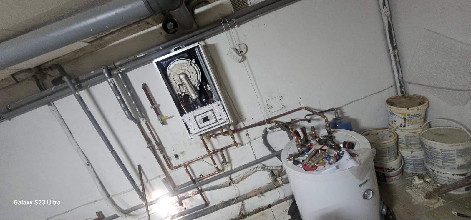 Montaż pieca gazowego z widoczną instalacją hydrauliczną i zbiornikiem. Rury miedziane prowadzone wzdłuż ściany. Widoczne puszki z farbą. Ujęcie z dołu.