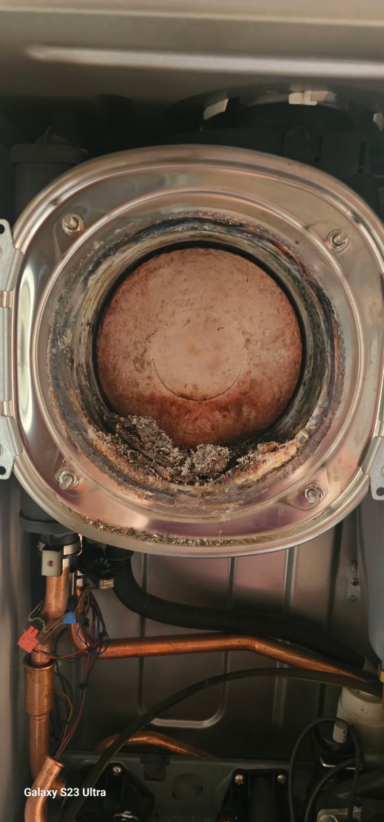 Zanieczyszczony palnik pieca gazowego z widocznymi osadami i korozją. Miedziane rurki instalacji hydraulicznej w dolnej części kadru. Wnętrze pieca w zbliżeniu.